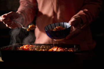 Close-up of a person plating a warm, steamy dish into a blue-striped bowl, focusing on the...