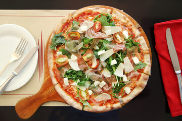A flat-lay view of a pizza topped with arugula, prosciutto, cherry tomatoes, and shaved parmesan on a wooden serving board