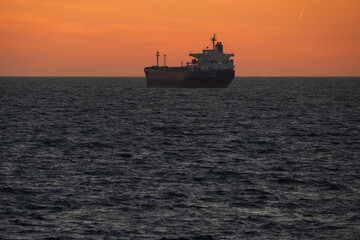 A cargo ship majestically sails on a calm sea against the backdrop of a stunning orange sunset,...