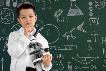 Thoughtful little boy with microscope near chalkboard at chemistry lesson in classroom