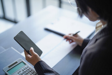 Asian businesswoman in suit holding smartphone and using pen writing on documents