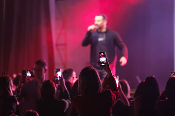 Audience in front of stage using mobile devices to record live event performance under colorful lighting.