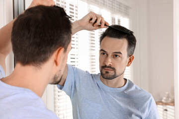 Fototapeta premium Handsome man styling his hair with comb near mirror at home