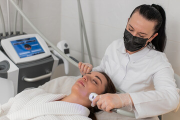 Caucasian girl lies with eyes closed while cosmetologist performs RF lifting vacuum facial massage....