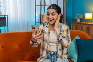 Indian young woman at home sitting on sofa with mobile phone joyfully screaming, celebrating...
