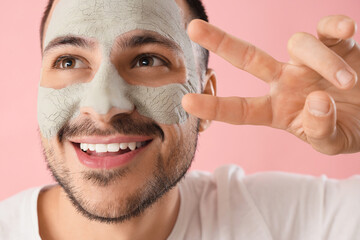 Handsome young happy man with cosmetic clay mask on face showing victory gesture against pink...