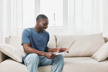 Happy young African man using laptop while sitting on sofa, enjoying online video or chatting at...