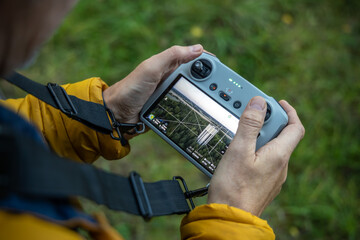 Close-up of a drone remote controller held in hands, showing the screen with a live view of a water tower and surrounding forest from the drone camera during flight.