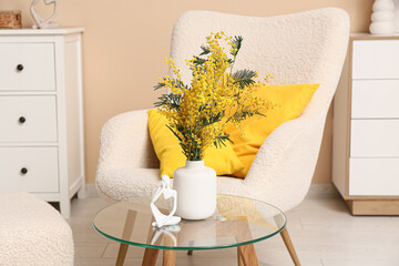 Vase with mimosa flowers and statuette on table near armchair in living room