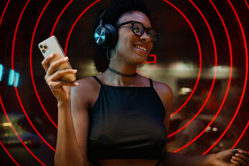 Stylish young woman with headphones and smartphone enjoying online music at night, smiling joyfully near glowing red light circles, fully immersed in her favorite songs