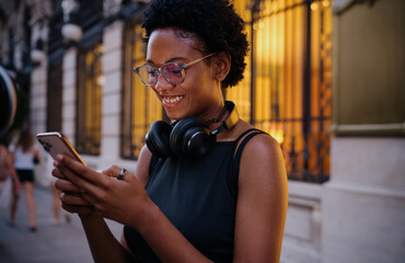 Cheerful Black woman walking down city street while smiling at her phone, using mobile app for...