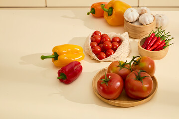 Tomatoes on a round wooden plate next to a bag of cherry tomatoes. Wooden bowls containing garlic and chili peppers. Fresh bell peppers with shiny peels. Shadows of objects cast on the kitchen table.
