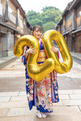 On a summer day, a 20-year-old Japanese girl wearing a blue kimono is in the traditional townscape of a Japanese city. She is holding large golden balloons shaped like the numbers 2 and 0.