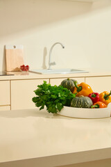 Celery, pumpkin, and bell peppers are placed on a tall white round plate on a marble countertop in the kitchen. Behind is a kitchen sink, wooden cutting board and a glass bowl of cherry tomatoes.