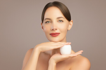 Beauty portrait of young elegant woman with sleek brown hair and glowing skin holding a white cream...