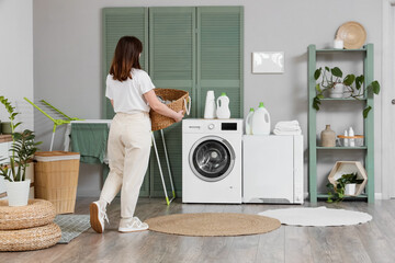 Young woman with wicker basket walking towards modern washing machine in laundry room