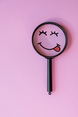 Vertical photo. A smiley face with a smile and tongue sticking out, drawn inside a magnifying glass on a pink background. The concept of search, the search for happiness and positive emotions.