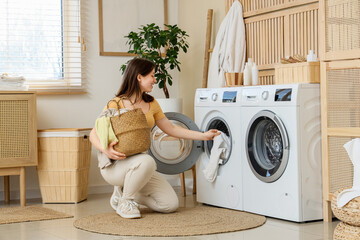 Young woman putting clothes into modern washing machine in laundry room