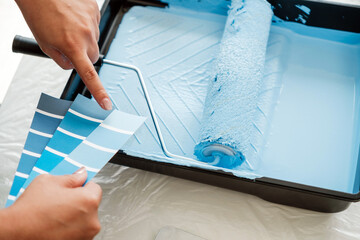 Close-up of a woman comparing blue paint swatches with a loaded roller in a paint tray during a...