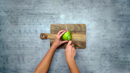 Close-up of a woman hands holding a knife cutting a green fresh apple on a wooden cutting board, with a gray background blank space.