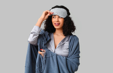 Beautiful young African-American woman in pajamas with cozy blanket and sleep mask on grey...