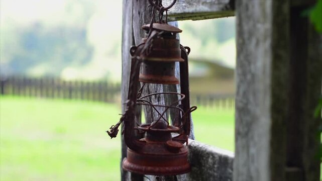 Stylish close up shot of an old rusty paraffin lamp hanging on a wooden gate, with an open paddock and fence in bokeh in the background