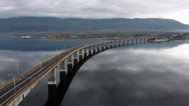 wide aerial View of High Bridge of Servia at lake polyfytos Kozani greece while tracking following cars , we see the reflection of the clouds at the lake and the bridge at the middle of the frame