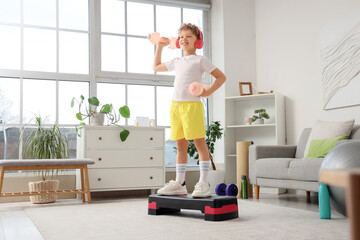 Sporty little boy in headphones exercising with dumbbells on aerobic stepper at home