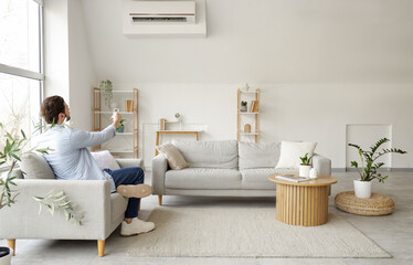 Young man turning air conditioner on sofa in living room, back view