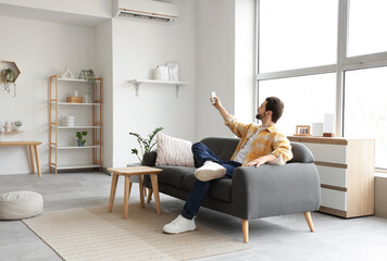 Young man turning on air conditioner from sofa at home