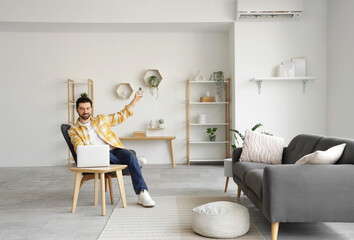 Young man with laptop in armchair turning on air conditioner at home
