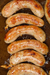 Several plump homemade sausages are sizzling in a dark frying pan, browning nicely in their own rendered fat