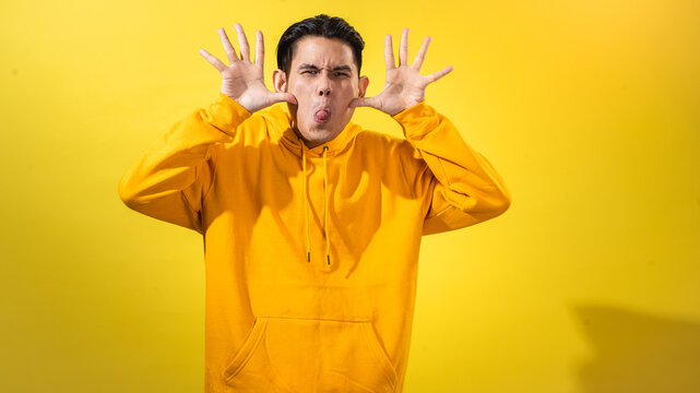 Asian man in yellow hoodie making a silly face by pulling cheeks and sticking out tongue, with both hands raised against a bright yellow background. Playful, comedic and humorous expression