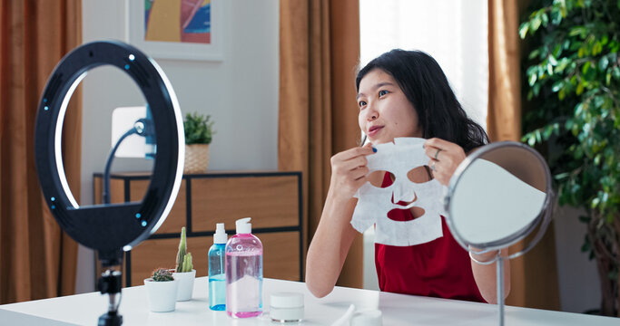 Beauty influencer films skincare vlog while gently removing makeup with cotton pad. She watches herself in the mirror and uses ring light and smartphone to capture clear footage.