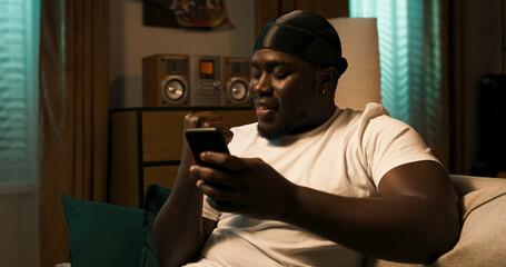 Relaxed man with a phone in hand sits on the couch, checking apps, browsing online stores, and planning purchases while adding notes and tasks to his calendar.