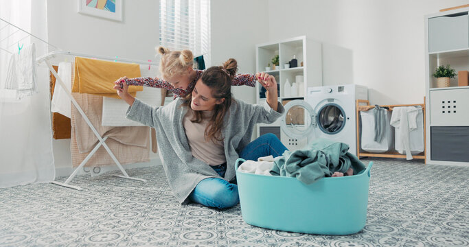 A young mother folds laundry while her daughter climbs on her back. Together they smile as the woman stretches the girl's arms wide like an airplane.