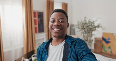 A cheerful male worker films a video for social media in his rented flat. He smiles, shows packed...
