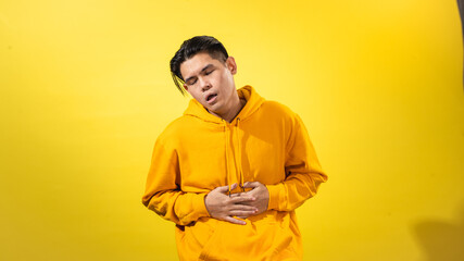 Sick young Asian man in yellow hoodie clutching his stomach with a painful expression, eyes closed,...