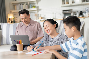 Asian grandmother helping grandson doing homework while grandfather working with laptop at home. Family spending time on weekend together