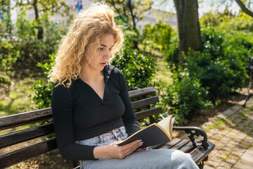 Obraz premium Young beautiful girl with curly hair sitting on a park bench, enjoying the nice weather while reading an interesting book. University student reading one more time before final exam at college.