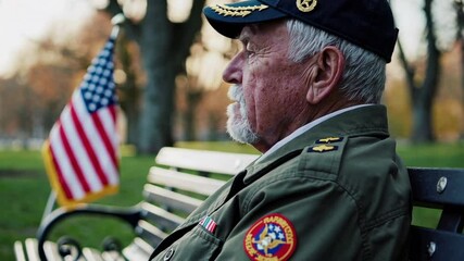 Elderly veteran in uniform sits on a bench, contemplating near a waving american flag in a peaceful park setting, honoring his service and sacrifice - Powered by Adobe