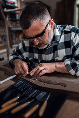 Carpenter sitting in his workshop restoring old piece of furniture with carving tool.