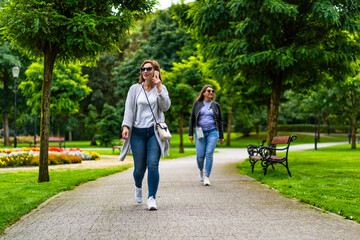 Obraz premium Two women of european beauty walking separately, one going in front and talking on phone, other in back, in city park 