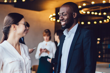 Successful male and female partners enjoying live communication during working time in office interior, diverse colleagues discussing business experience standing in company workspace and smiling