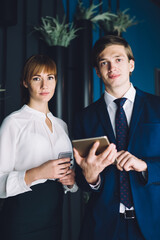Half length portrait of confident male and female colleagues holding digital devices and looking at camera during office meeting, Caucasian managers in formal clothing posing with technology in hands