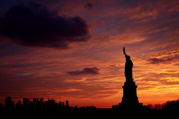 Naklejka premium Silhouette of Statue Liberty against fiery sunset sky, cityscape background. Represents freedom, hope, and American identity