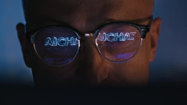 Male AI chat bot developer working on a computer in a futuristic research laboratory. Computer screen reflecting in eyeglasses displaying text "AI Chatbot"