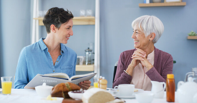 Elderly woman, caregiver and reading book in home, breakfast or smile in retirement in dining room. People, connection and literature in morning for care, storytelling and support with novel in house