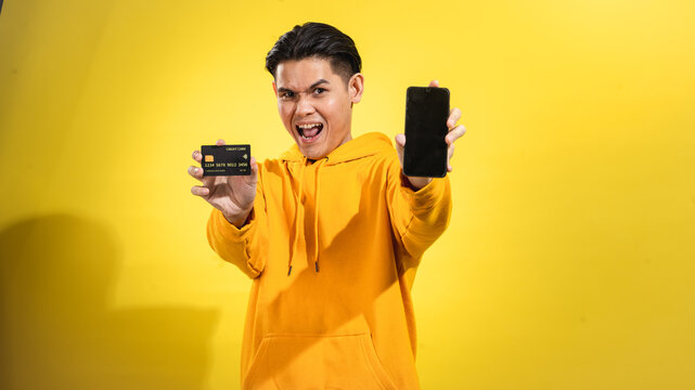 Excited young Asian man in yellow hoodie holding a credit card and smartphone toward the camera, smiling against a solid yellow background. Studio concept of mobile banking and online payments