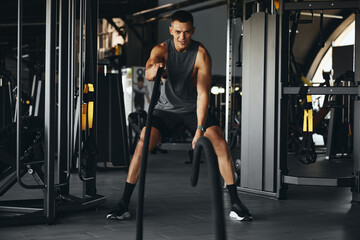 Powerful and muscular male in sportswear using battle ropes for exercise at fitness center. Athlete working out with battle ropes in a health club. Bodybuilding, fitness, and health concept
